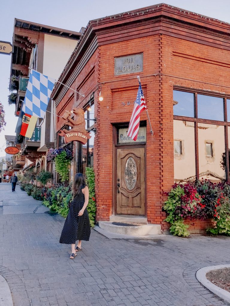 girl with long dark hair wearing a black and white polka dotted maxi dress strolling in front of a red brick building with flags on it in Leavenworth, Washington. Colorful Bavarian style buildings in the background with pretty colorful flowers blooming everywhere.