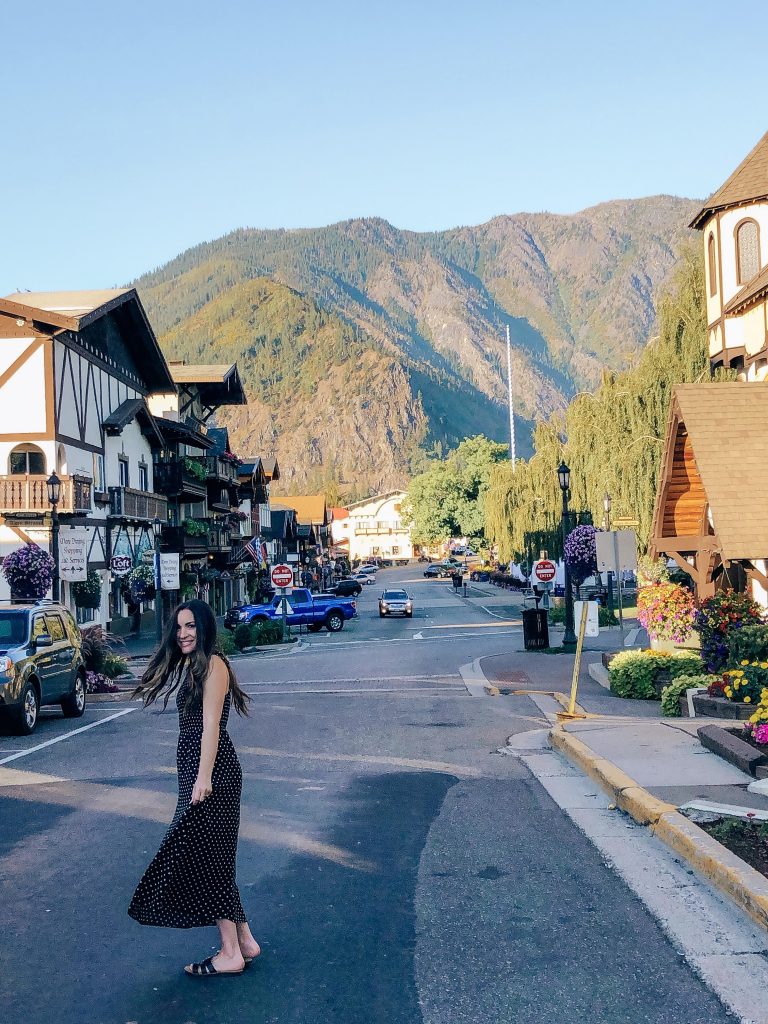 girl with long dark hair wearing a black and white polka dotted maxi dress strolling down Front Street in Leavenworth, Washington. Colorful Bavarian style buildings in the background with pretty colorful flowers blooming everywhere. Beautiful view of the Cascade mountains in the background.